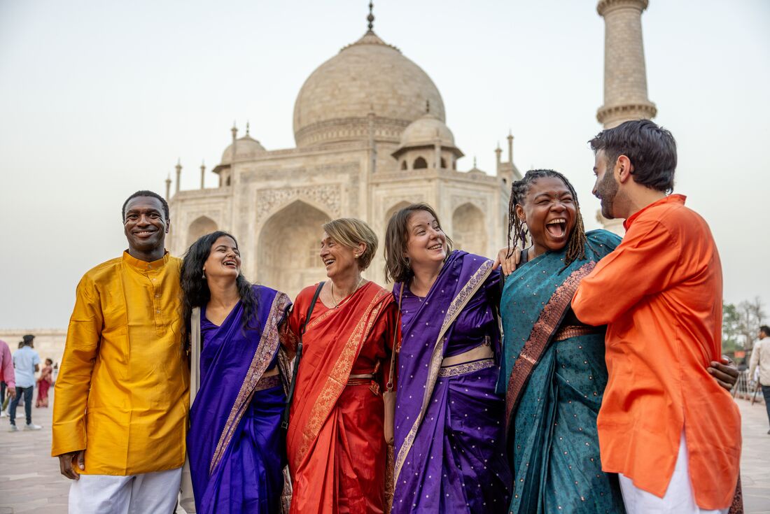 Travellers in opulent colourful garments smile and laugh happily together outside the Taj Mahal at sunset