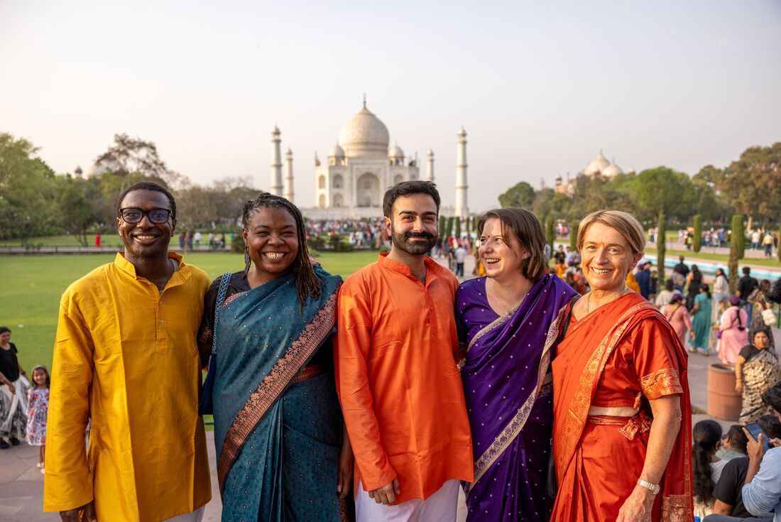 Group of varied ages pose with smiles in colourful elegant clothing standing at a distance in front of the Taj Mahal
