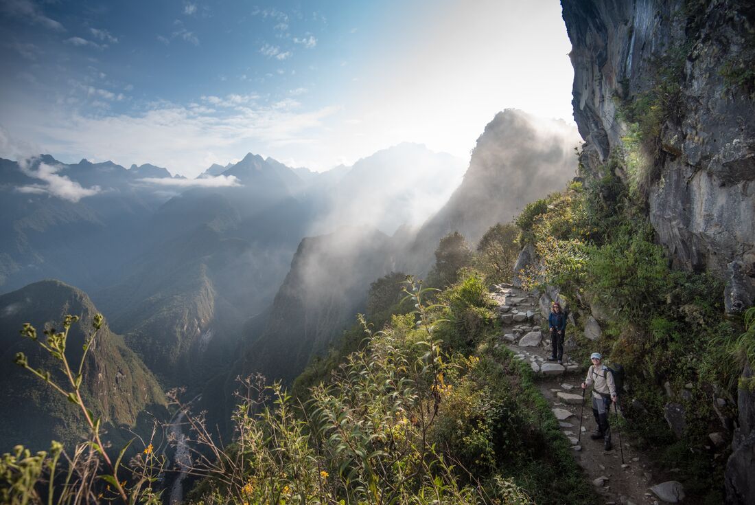 Travellers look back at the photographer as sunlight hits a gentle mist around the bend of a mountain path on the Inca Trail