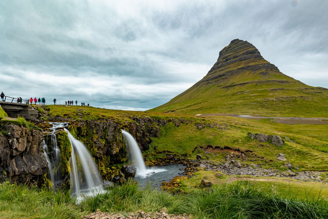 Wide shot of crowds watching a pair of waterfalls fall off a short cliff flowing into a creek with large mountain in background